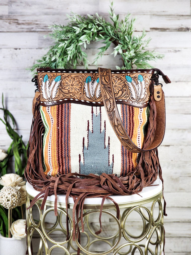 Concealed carry fringe handbag with Southwestern saddle blanket made from woven rug. Top leather paneling is tooled with floral and painted feathers. Fringe hangs from side of bag with tooled feather shoulder strap visible. Purse is displayed on marble table. 