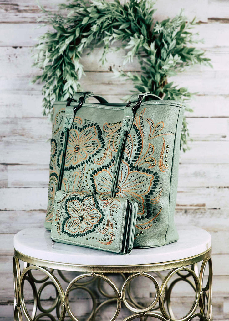Concealed carry green tote with think green whip stitch outlining the orange embroidered  floral patterns with silver accent studs. Strap is decorated with "X" whip stitch and fringe. Matching wallet lays next to handbag. Angled on a marble stool. 