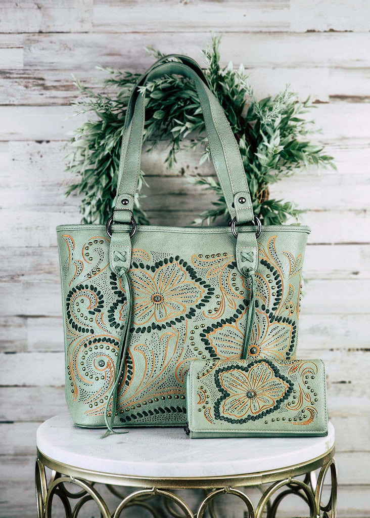 Concealed carry green tote with think green whip stitch outlining the orange embroidered  floral patterns with silver accent studs. Strap is decorated with "X" whip stitch and fringe. Matching wallet lays next to handbag. Handbag and wallet set sits on marble stool. 