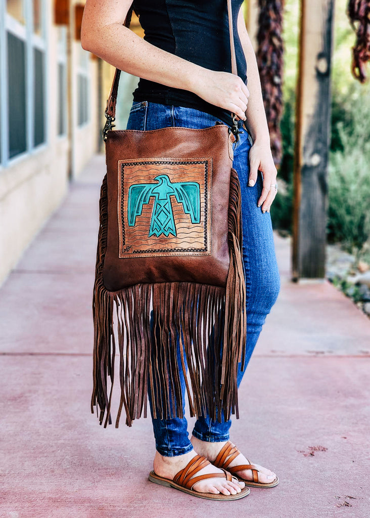 Medium oil concealed carry crossbody with a tooled thunderbird add on leather. Fringe hangs from around the purse. Model wears bag with shoulder strap.