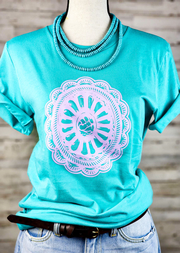 Teal short sleeve tee with lavender print concho design.  Shirt hangs from mannequin with turquoise like stone choker. 