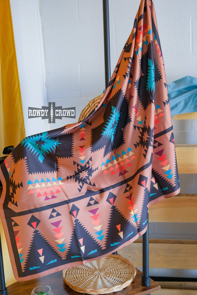 Clay brown wild rag with southwestern design with multiple colors. Rag is outlined with a black border before design starts in the middle. Rag hangs from metal stand. 