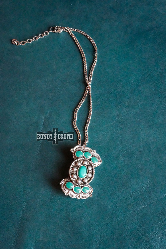 Turquoise like stone necklace with antique silver concho design. Pendant is on a chain and is displayed on a turquoise fabric background. 