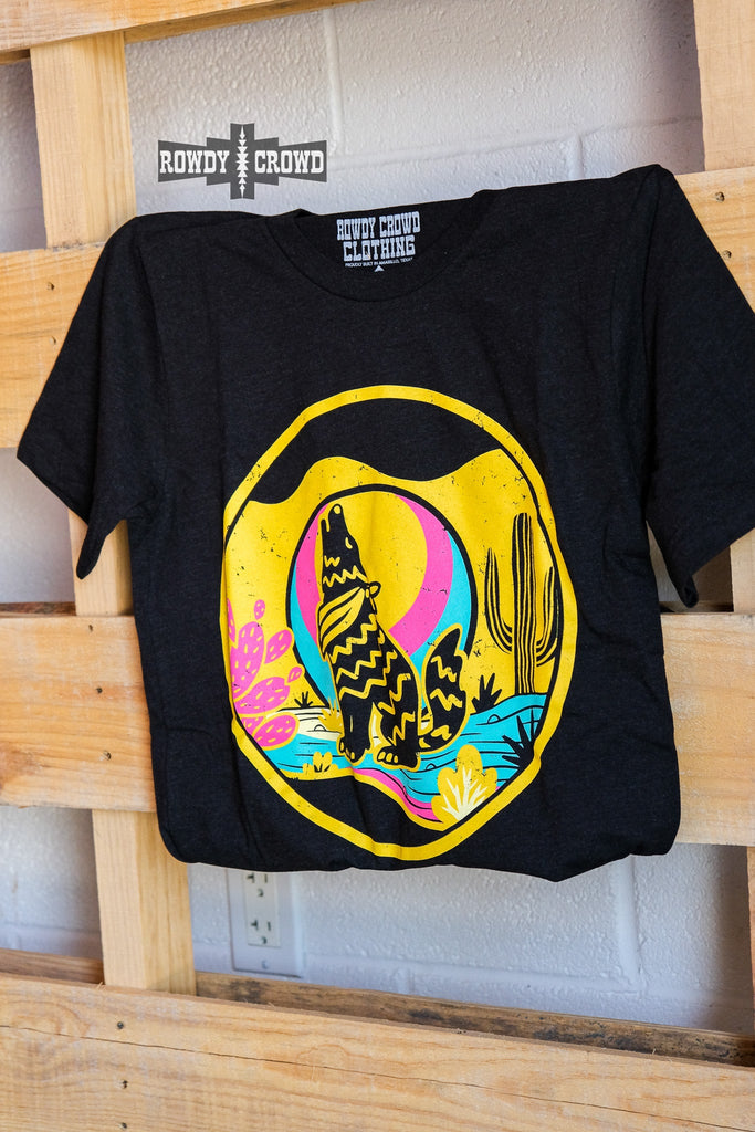Heather black short sleeve tee with a yellow, blue, and pink desert scene of a coyote howling at the moon. Tee hangs from a wooden pallet. 