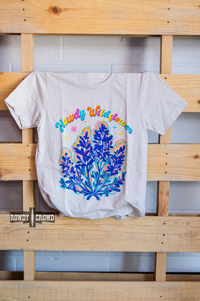 Natural short sleeve with colorful bubble lettering reading "Howdy Wildflower". Wildflower colorful graphic with white heart designs. Tee hangs from wooden pallet.
