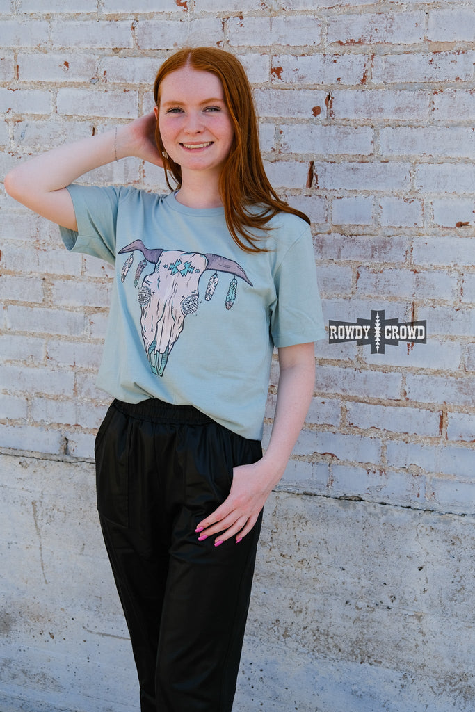 Teal short sleeve tee with a cow skull with disco eyes and feathers hanging from the steer horns. Model wears this with black joggers. 