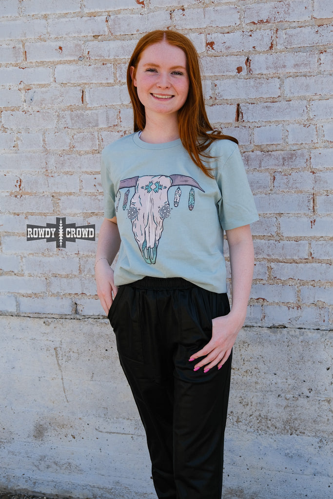 Teal short sleeve tee with a cow skull with disco eyes and feathers hanging from the steer horns. Model wears this with black joggers. 