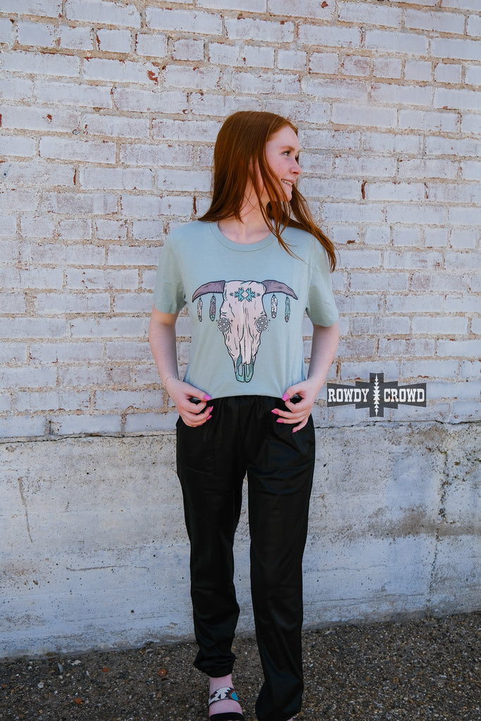 Teal short sleeve tee with a cow skull with disco eyes and feathers hanging from the steer horns. Model wears this with black joggers against a white brick wall. 