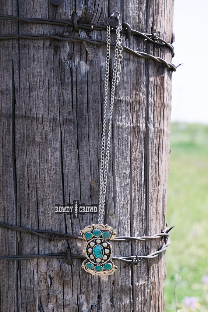 Turquoise like stone necklace with antique silver concho design. Pendant is on a chain and is displayed on a wooden fence post with barbed wire. 