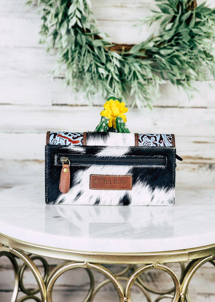 Cowhide wallet with blue embossed floral leather panels on the flap of the wallet. Wallet sits on a marble stool. Backside of the wallet zippered coin pocket shows. 