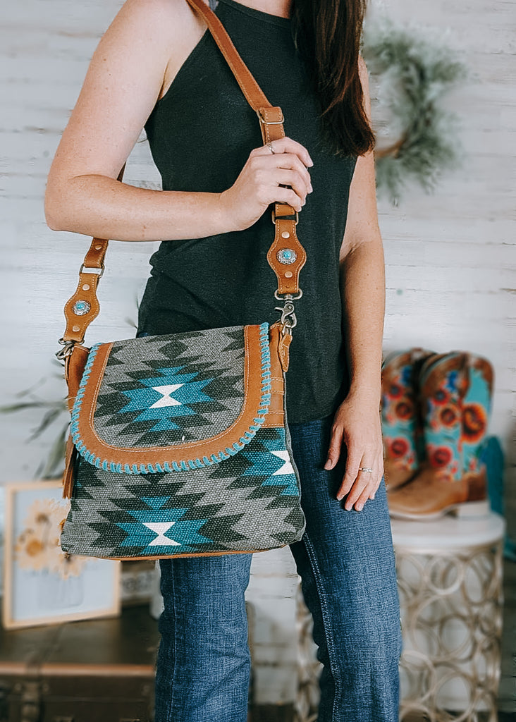 Gray & Turquoise Aztec Shoulder Handbag on model with cute western decor farmhouse in background including cowboy boots and box decor. Model is wearing a black tank and bootcut jeans with handbag draped over.