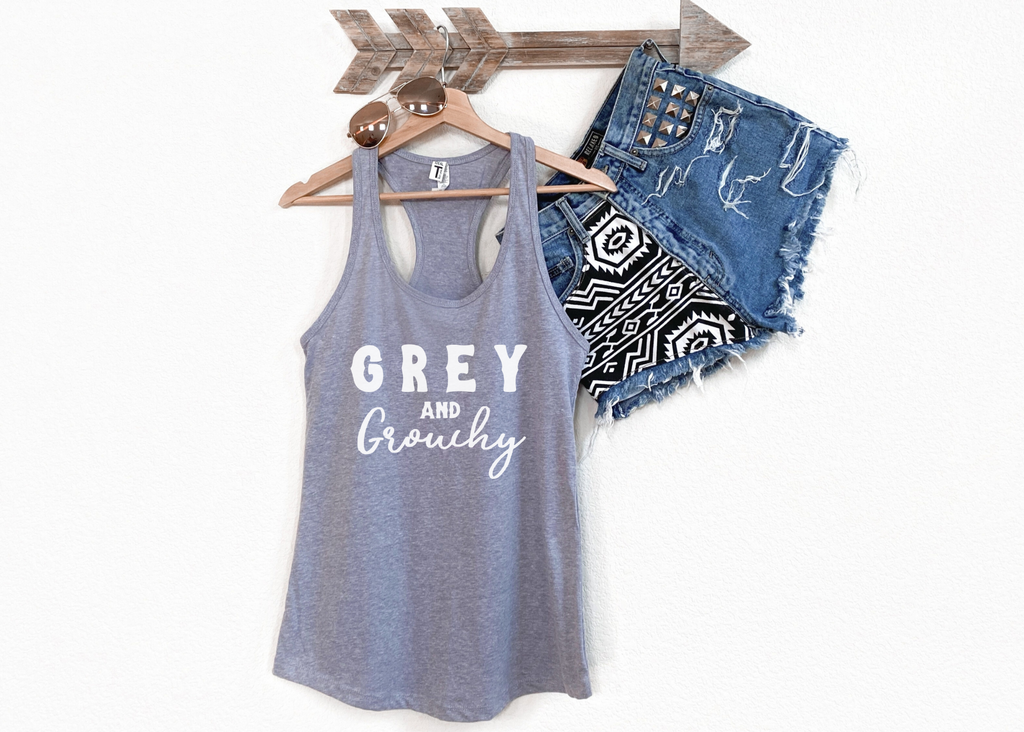 Grey tank top with white lettering reading "Grey and Grouchy". Tank hangs from wooden hanger with jean shorts.
