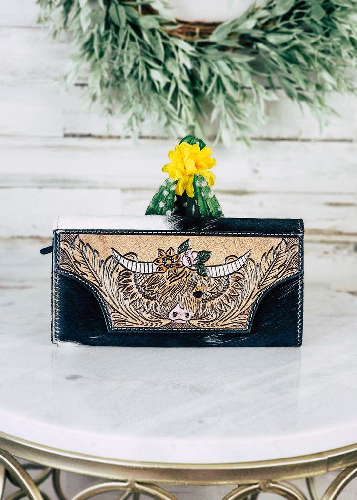 Cowhide wallet with tooled feathers surrounding a high land cow with sunflowers. Painted floral tooling accents are on top of the highland. Wallet sits on top if a marble table. 