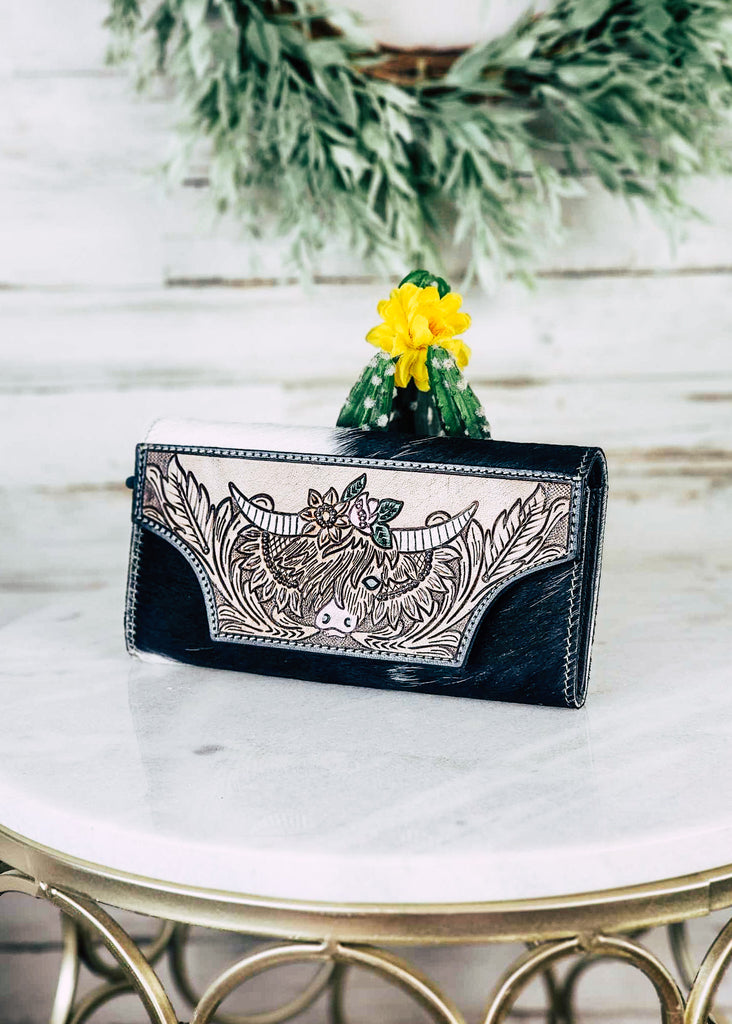 Cowhide wallet with tooled feathers surrounding a high land cow with sunflowers. Painted floral tooling accents are on top of the highland. Wallet sits on top if a marble table. 