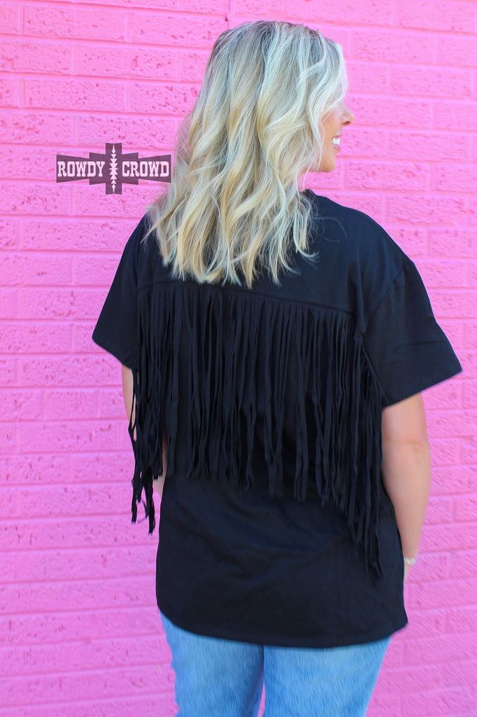 Western black short sleeve tee. Model is showing the back of the shirt which shows off the fringe. Model faces pink brick wall. 