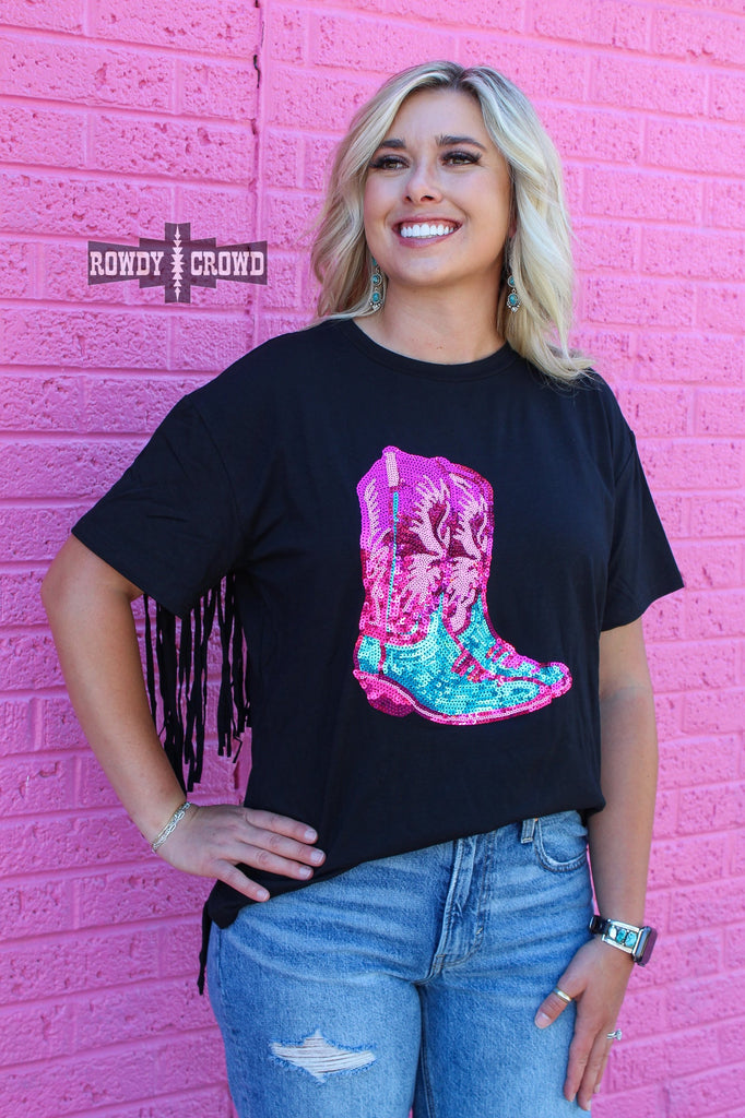 Black short sleeve tee with with a pink and turquoise sequin boot design on front of the tee. Fringe hangs on back of sleeve. Model is wearing distressed jeans and stand against a pink brick background. 