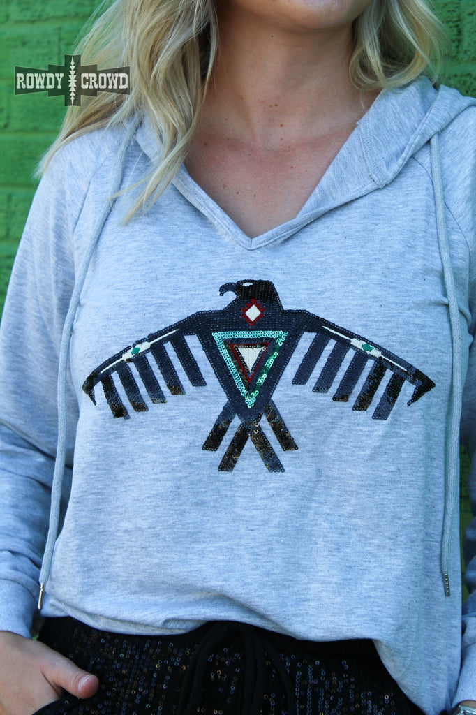 Grey hoodie with a thunderbird sequin in black with accents of turquoise and deep red. Model wears this with sparkle black pants against a green brick wall. Zoomed in on the design.