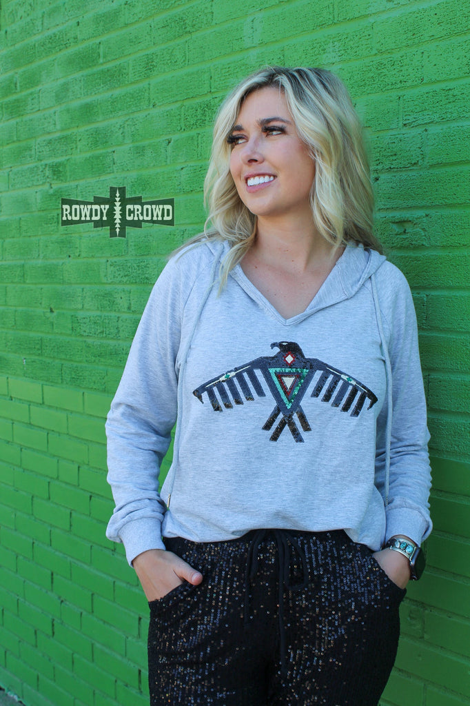 Grey hoodie with a thunderbird sequin in black with accents of turquoise and deep red. Model wears this with sparkle black pants against a green brick wall.