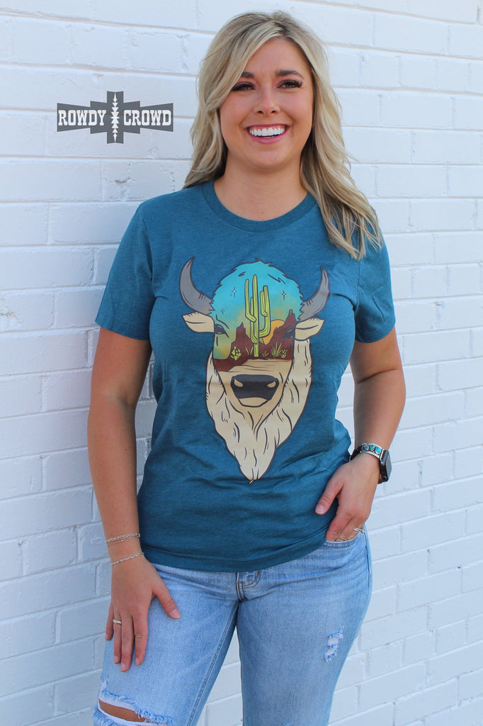 Teal short sleeve tee with a printed buffalo design with a desert scenery on the face. Model wears tee with distressed jeans against white brick. 