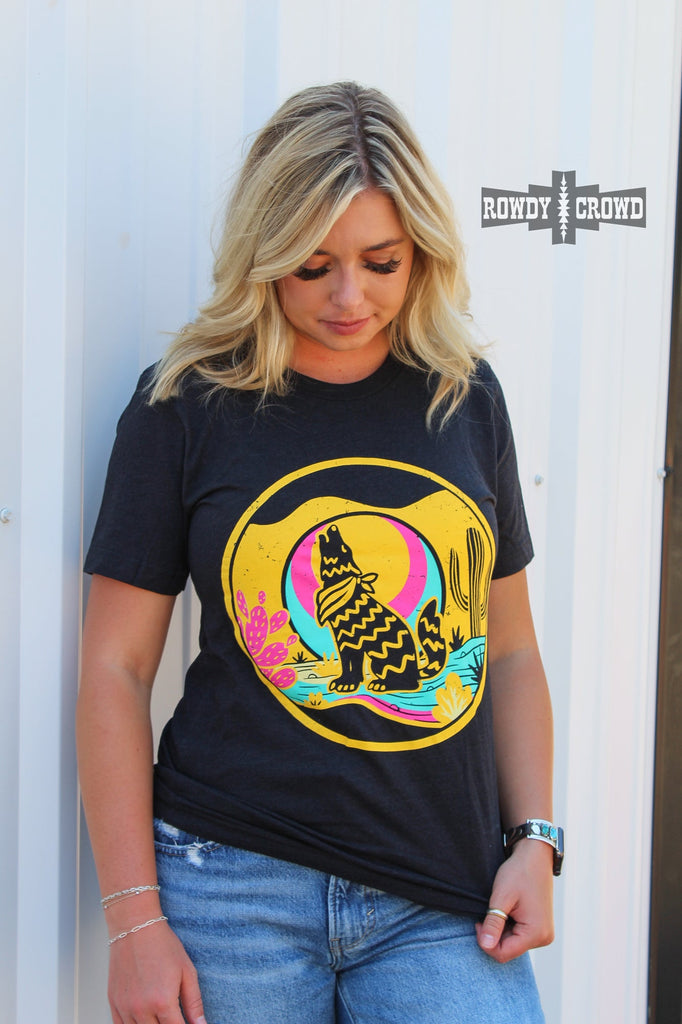 Heather black short sleeve tee with a yellow, blue, and pink desert scene of a coyote howling at the moon. Model wears this with jeans against a white background. 