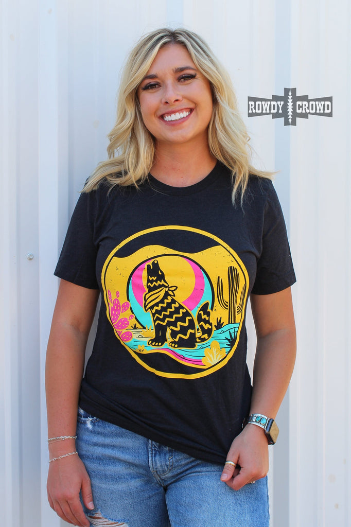 Heather black short sleeve tee with a yellow, blue, and pink desert scene of a coyote howling at the moon. Model wears this with distressed jeans with white background.