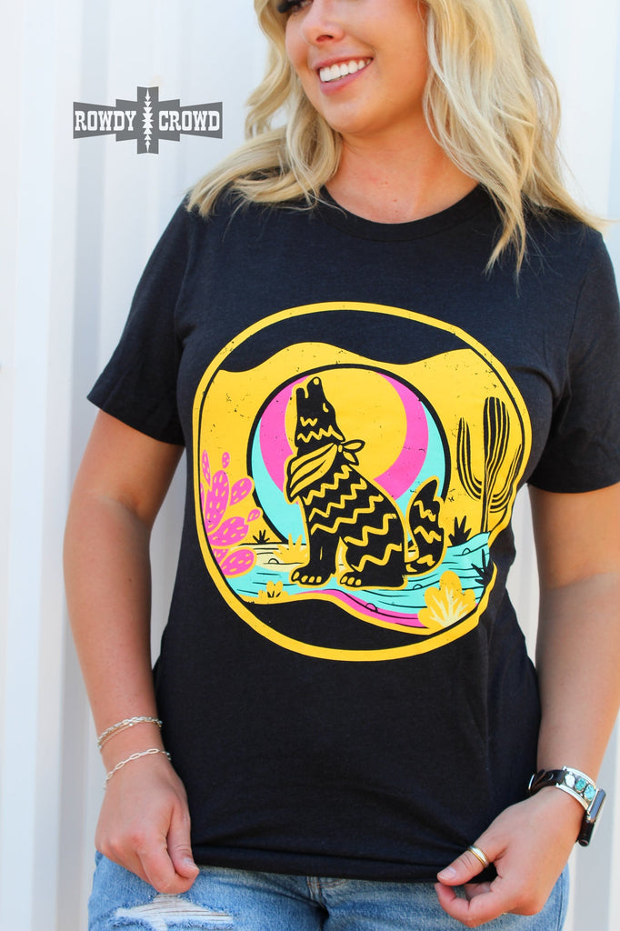 Heather black short sleeve tee with a yellow, blue, and pink desert scene of a coyote howling at the moon.  Model stretches out the tee showing the design better. 