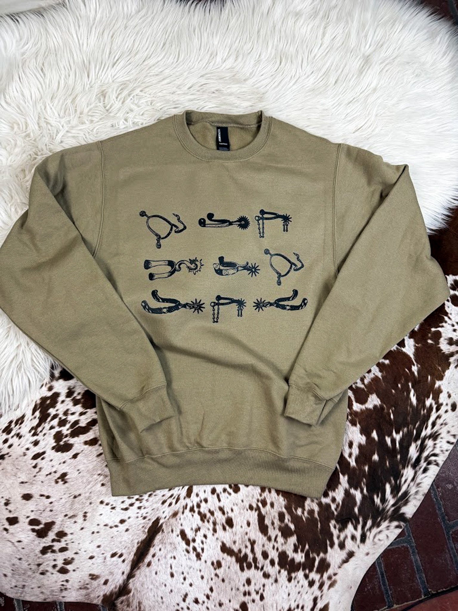Anbrown crewneck sweatshirt with black western-themed graphic prints, laid flat on a white faux fur rug and a brown and white cowhide rug.