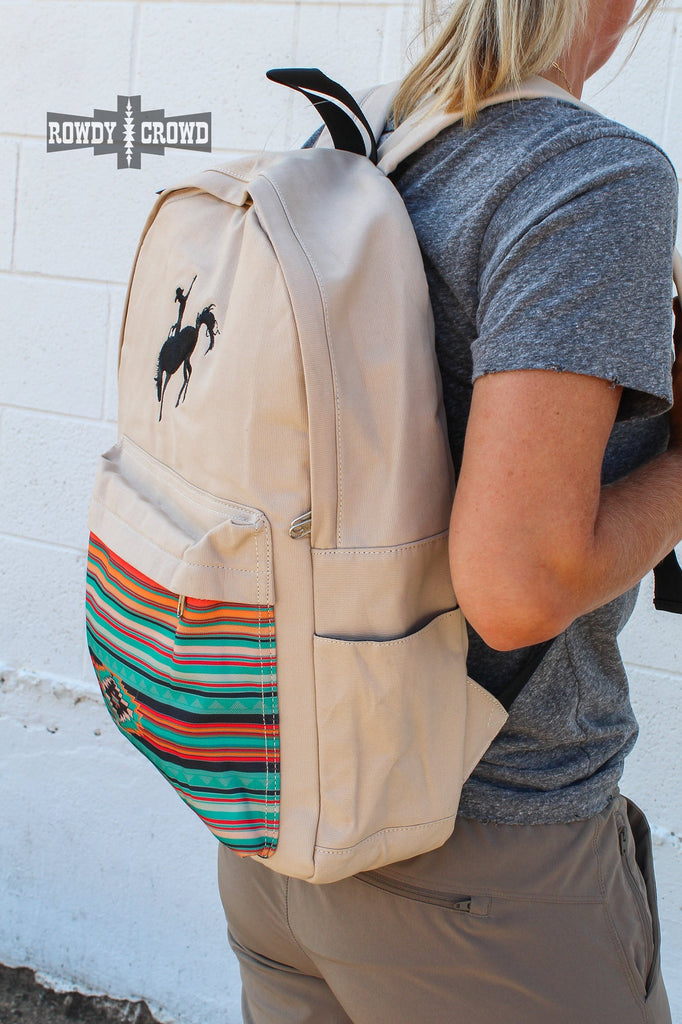 Tan canvas bag with black embroidered buckaroo. Front pocket has southwestern designed canvas accent. Side pockets are visible for water bottles.  Side angle of backpack on model with adjustable straps partly visible.