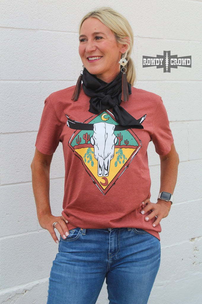 rust colored western graphic tee with steer skull image and cactus, model is blonde and wearing a wild rag with fringe earrings and denim jeans, white brick background