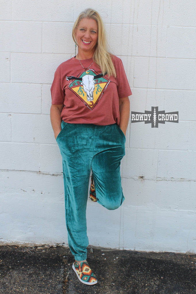 rust colored western graphic tee with steer skull image and cactus, model is blonde and wearing a wild rag with fringe earrings and turquoise joggers, white brick background
