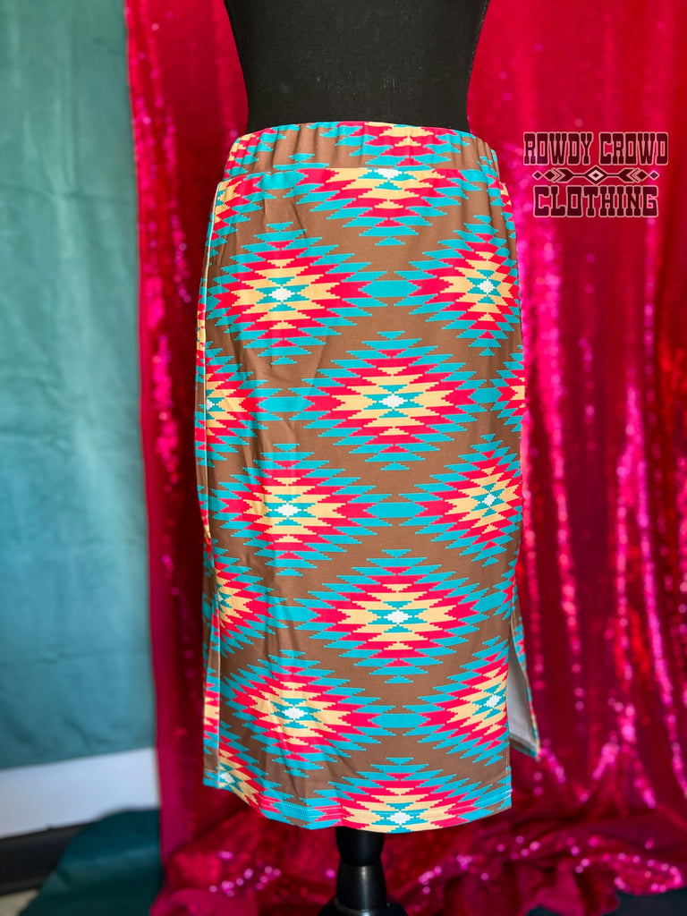 Southwestern tribal printed knee-length skirt with elastic band waist with slit up the side to mid-thigh. Brown is the main color with tribal design in bright teal, pink, and yellow. Hanging on mannequin. 