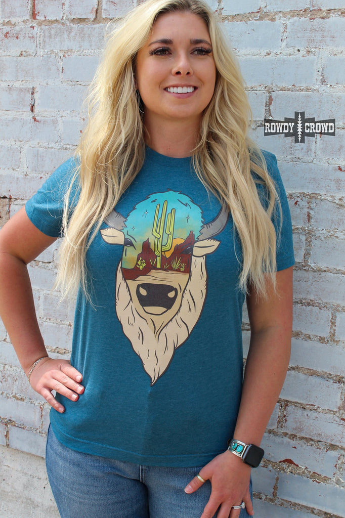 Teal short sleeve tee with a printed buffalo design with a desert scenery on the face. Model wears tee with distressed jeans against white brick. 