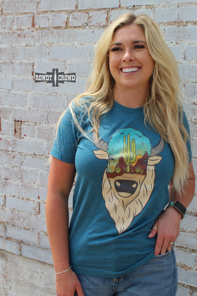 Teal short sleeve tee with a printed buffalo design with a desert scenery on the face. Model wears tee with distressed jeans against white brick. 