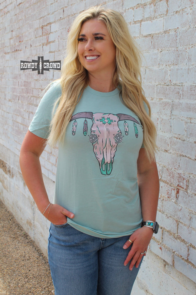 Teal short sleeve tee with a cow skull with disco eyes and feathers hanging from the steer horns. Model wears this with jeans against a white brick wall.