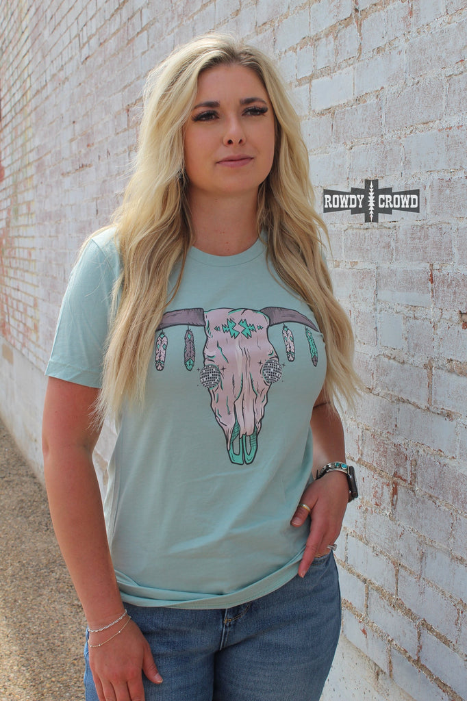 Teal short sleeve tee with a cow skull with disco eyes and feathers hanging from the steer horns. Model wears this with jeans against a brick wall. 