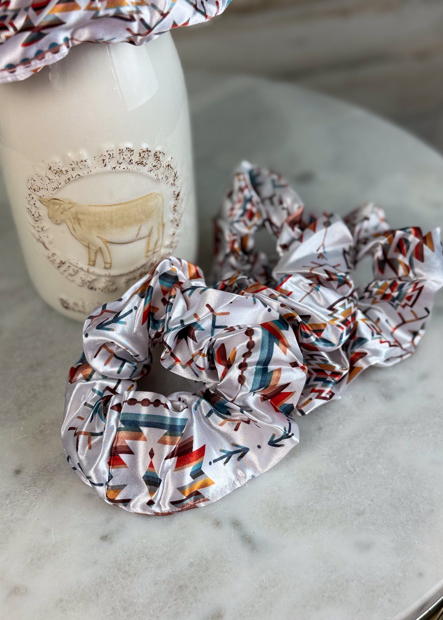 A set of shiny satin hair scrunchies with a Southwestern geometric pattern in orange, teal, brown, and black on a white base, displayed on a marble surface next to a white ceramic bottle.