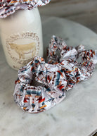 A set of shiny satin hair scrunchies with a Southwestern geometric pattern in orange, teal, brown, and black on a white base, displayed on a marble surface next to a white ceramic bottle.