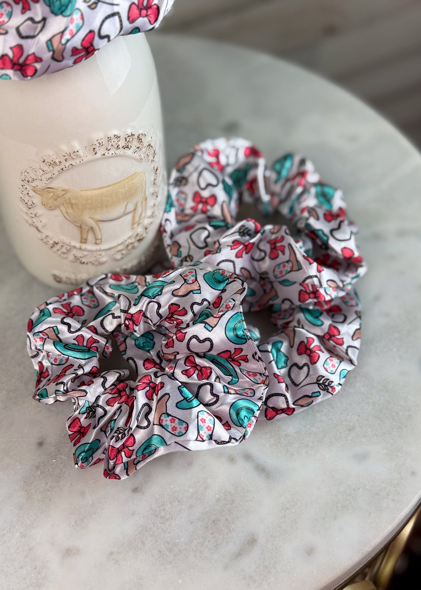 Two fabric hair scrunchies with a colorful cartoon pattern of hearts, bows, and cartoon characters on a white background, displayed on a marble surface next to a white ceramic bottle.