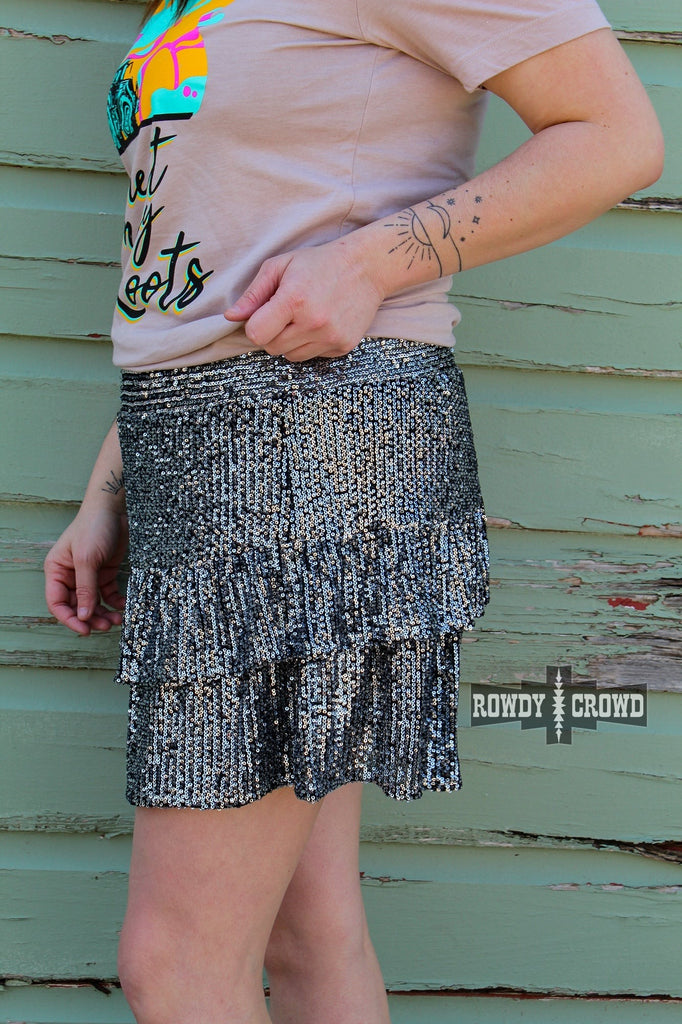 Black sequin shorts with ruffles on side profile model with pink shirt up against a green wood wall. 