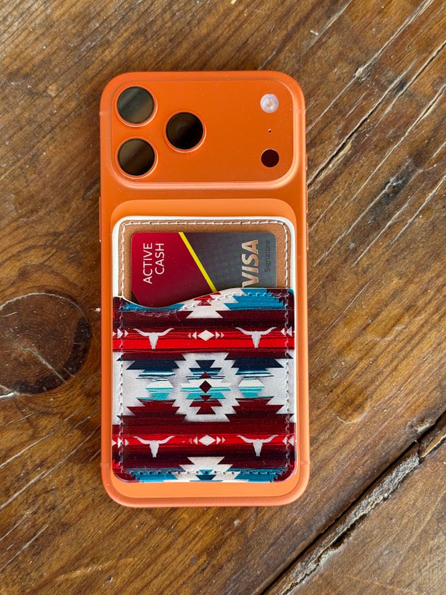 An orange protective phone case with a built-in card holder featuring a red, blue, and white Southwestern geometric pattern, shown on a rustic wooden surface.