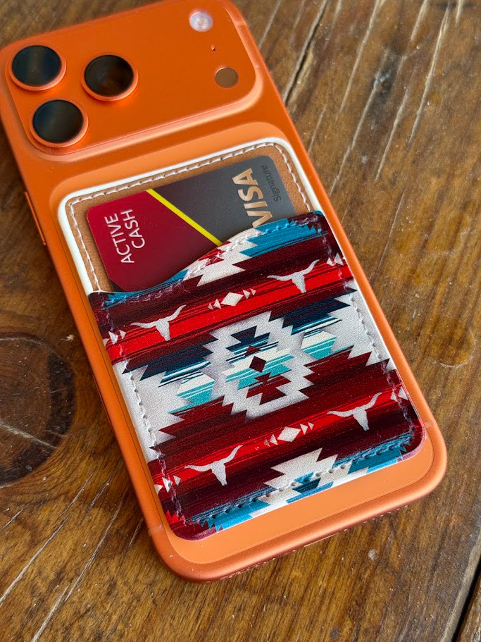An orange phone case with a western-style card holder pocket, shown on a wooden table. The pocket features a red, white, and blue geometric patterned leather insert holding two cards.