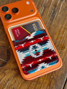 An orange phone case with a western-style card holder pocket, shown on a wooden table. The pocket features a red, white, and blue geometric patterned leather insert holding two cards.