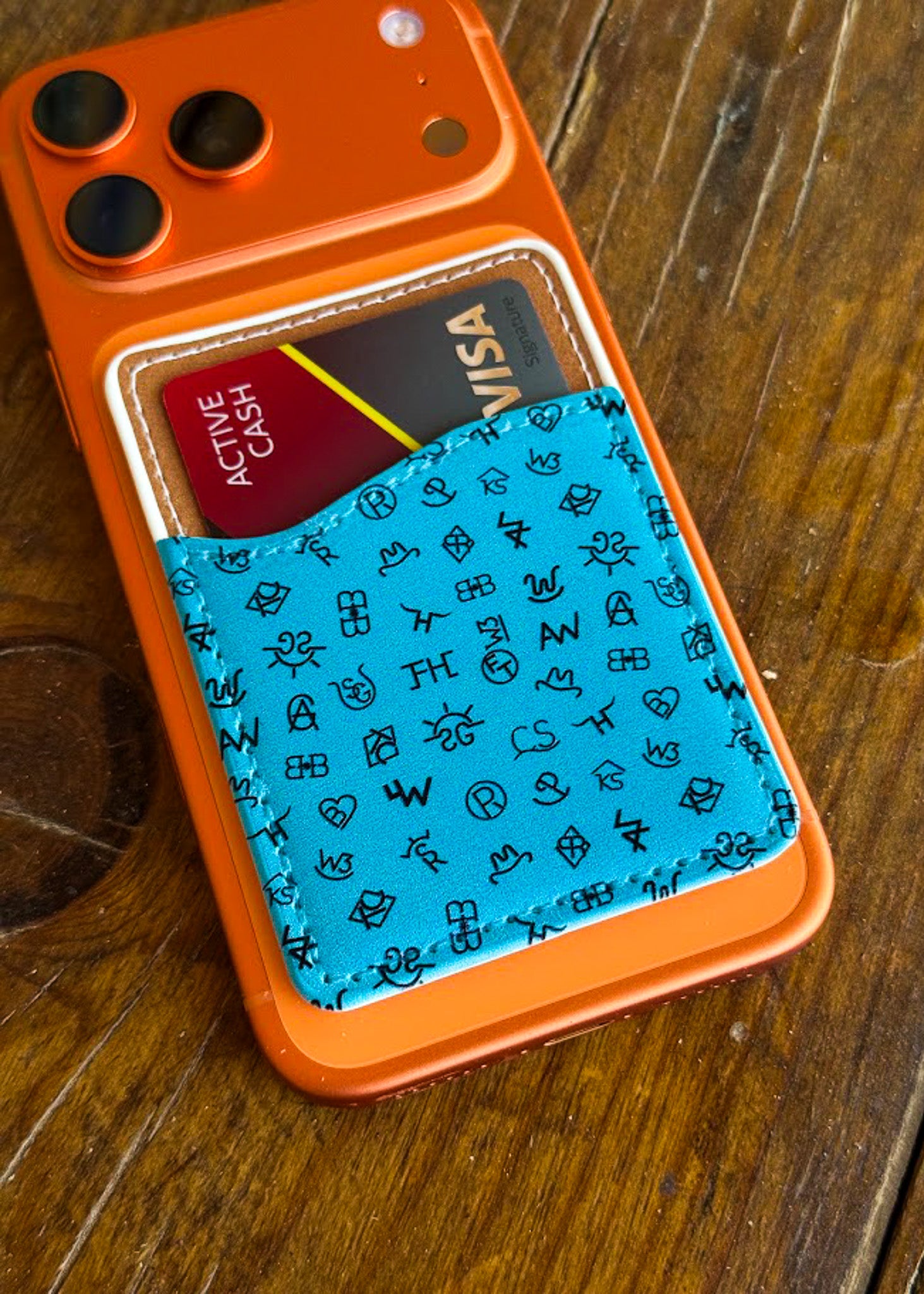An orange phone case with a built-in card holder pocket, shown on a wooden table. The pocket contains a turquoise blue patterned fabric card holder with black symbols, holding a Visa card and another card underneath.