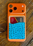 An orange phone case with a built-in card holder, shown on a rustic wooden surface. The lower half of the case features a bright blue textured panel decorated with small black hand-drawn style icons, and it holds two sample credit cards.