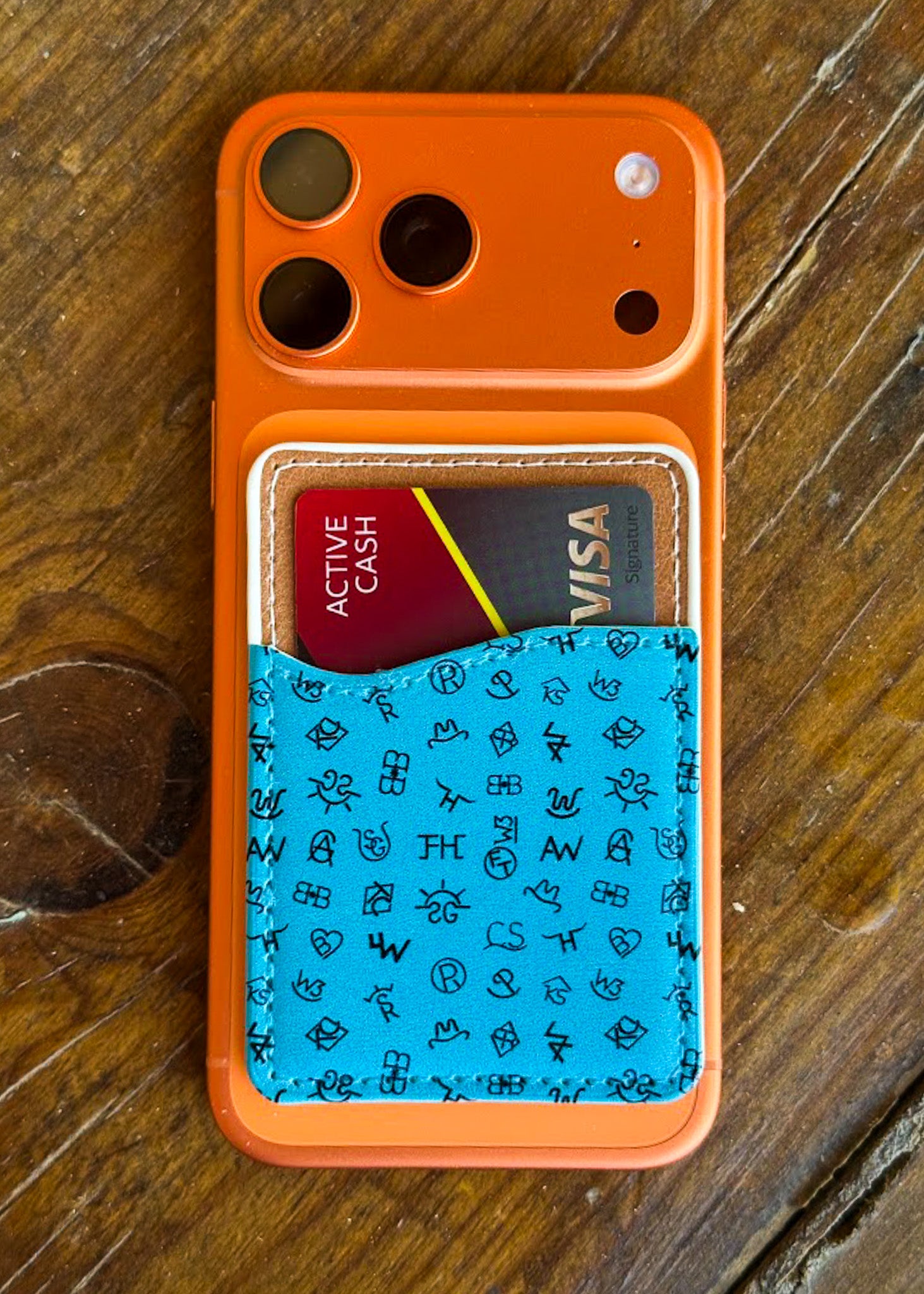 An orange phone case with a built-in card holder, shown on a rustic wooden surface. The lower half of the case features a bright blue textured panel decorated with small black hand-drawn style icons, and it holds two sample credit cards.