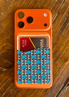 An orange phone case with a built-in card holder pocket, featuring a black, white, and turquoise patterned card sleeve, shown on a rustic wooden table.