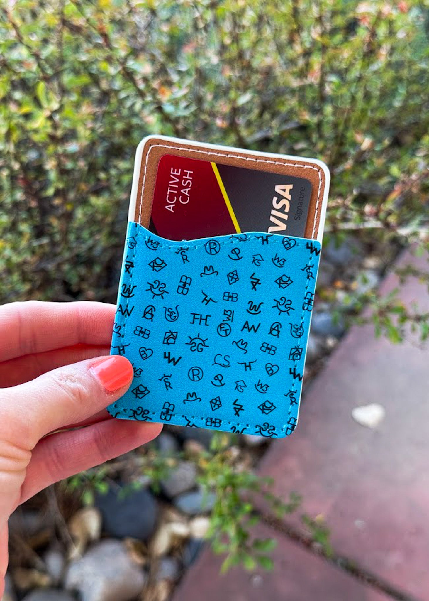 A hand holding a slim cardholder wallet with a turquoise blue patterned exterior and a brown interior, showing two cards partially inserted. The background is an outdoor blurred garden setting.