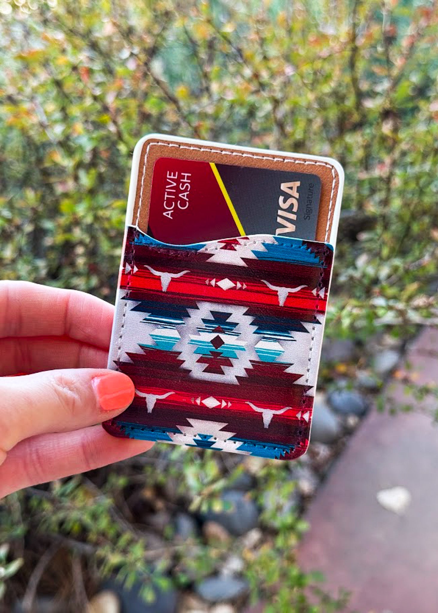 A hand with orange nail polish holds a slim cardholder wallet featuring a red, white, and blue Southwestern geometric pattern. The wallet contains credit cards, including one with a Visa logo.