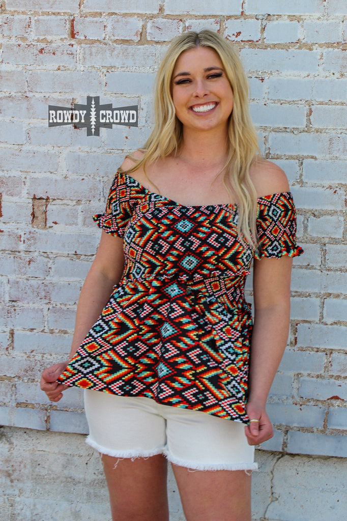 Southwestern design off the shoulder shirt. Bright southwestern colors inluded blue, red, greens, and more. Model with ruffle short sleeves stands against a white brick background. 