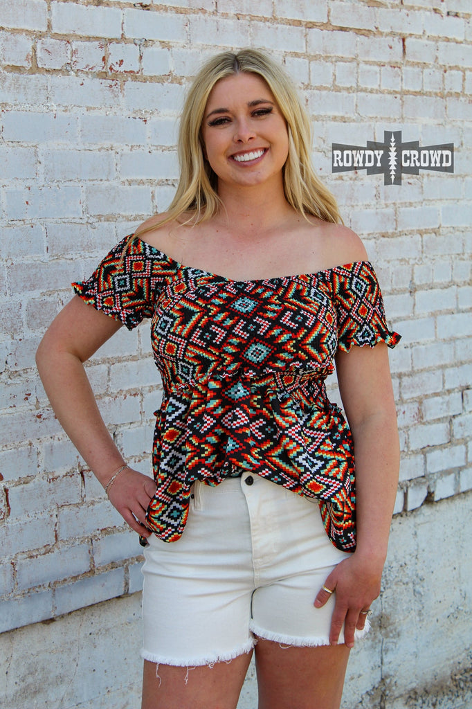 Southwestern design off the shoulder shirt. Bright southwestern colors inluded blue, red, greens, and more. Model provides side angle view with ruffle short sleeves. Part of shirt is tucked into shorts. 